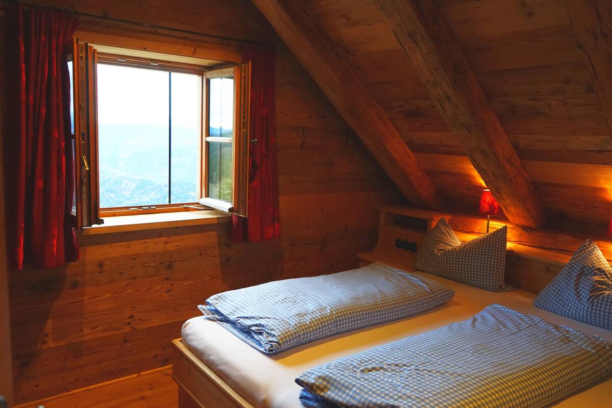 A wooden cabin room with a bed, a window with curtains, and a view of the mountains.