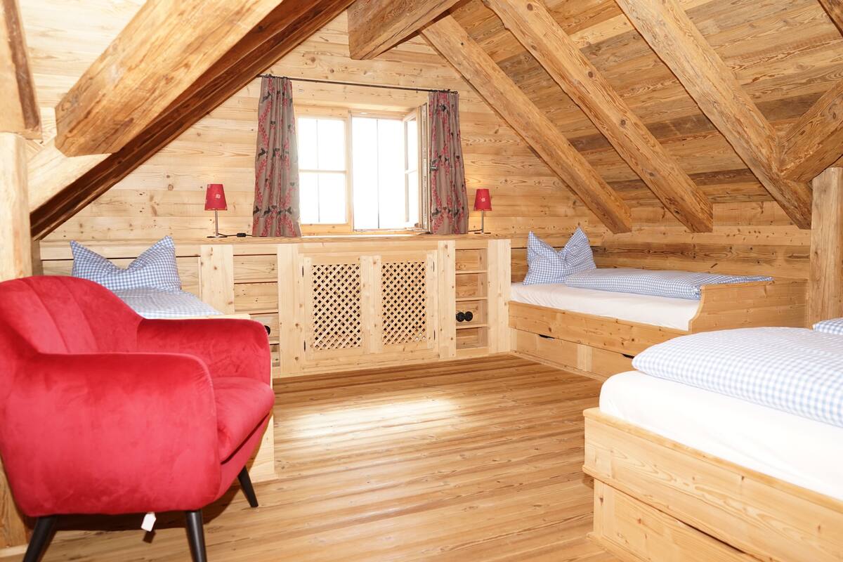 A wooden cabin with a loft bedroom, a red chair, and a desk with a lamp.