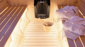 Una cabaña de madera con un ventanal circular que ofrece vistas a la montaña. En el interior, hay una cama con cabecera de listones de madera, una mesita de madera con un bol y una cuchara, y un bolso negro colgado en la pared.