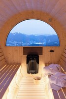 A wooden cabin with a circular window offering a mountain view. Inside, there is a bed with a wooden slat headboard, a small wooden table with a bowl and spoon, and a black bag hanging on the wall.