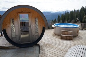 Estructura de madera con entrada circular, jacuzzi y terraza de madera con vistas a montañas.