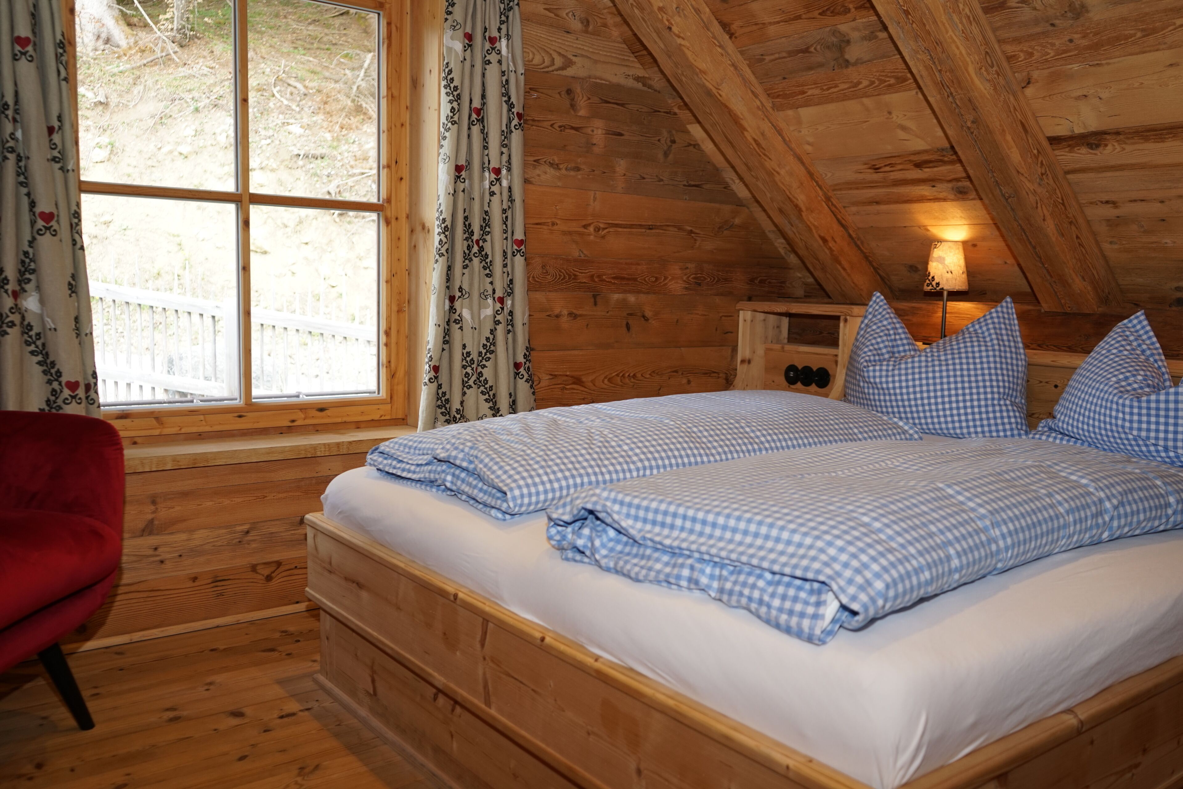 Un dormitorio de cabaña de madera con cama, una silla roja y una ventana con cortinas.