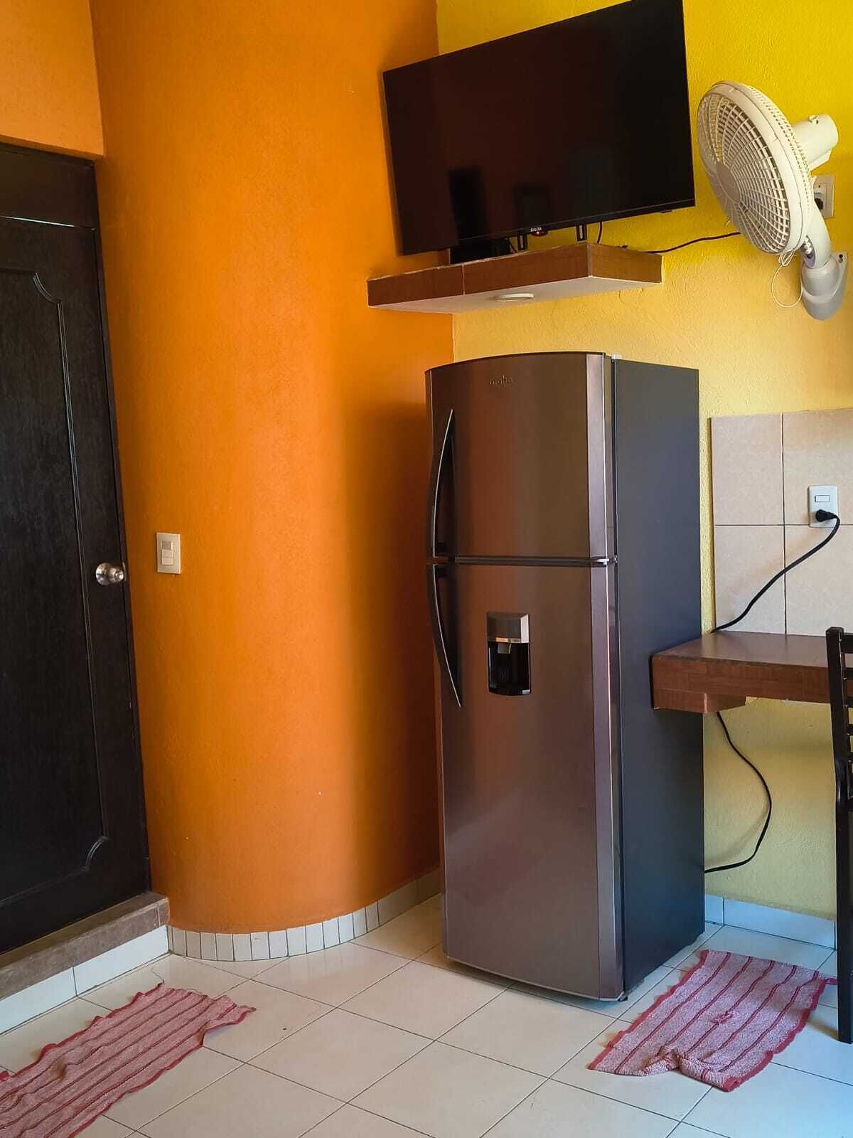 A kitchen with a refrigerator, a TV mounted on the wall, a fan, and a tiled floor.
