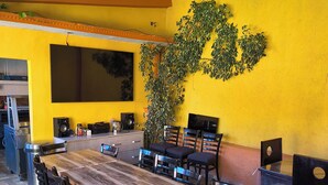 A dining area with a long wooden table, multiple chairs, a large potted plant, and a television mounted on the wall.