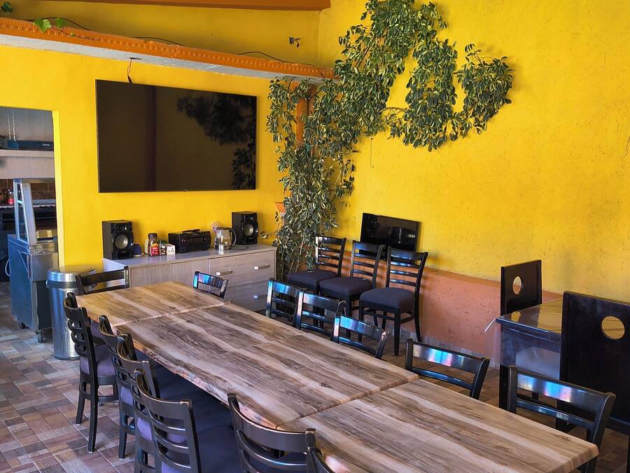 A dining area with a long wooden table, multiple chairs, a large potted plant, and a television mounted on the wall.