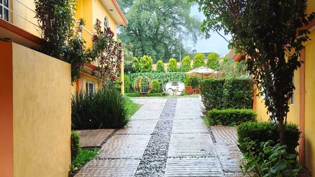 A pathway leading to a seating area with a small table and chairs, surrounded by greenery and a yellow building.