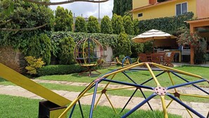 A garden with a swing set, a dining area with outdoor furniture, and a yellow building in the background.