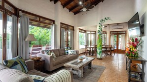 A spacious living area with a high ceiling, a sofa set, a dining table, and a TV mounted on the wall.