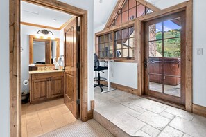 Un couloir avec une porte en bois donnant sur une salle de bain équipée d’un miroir et d’un lavabo.