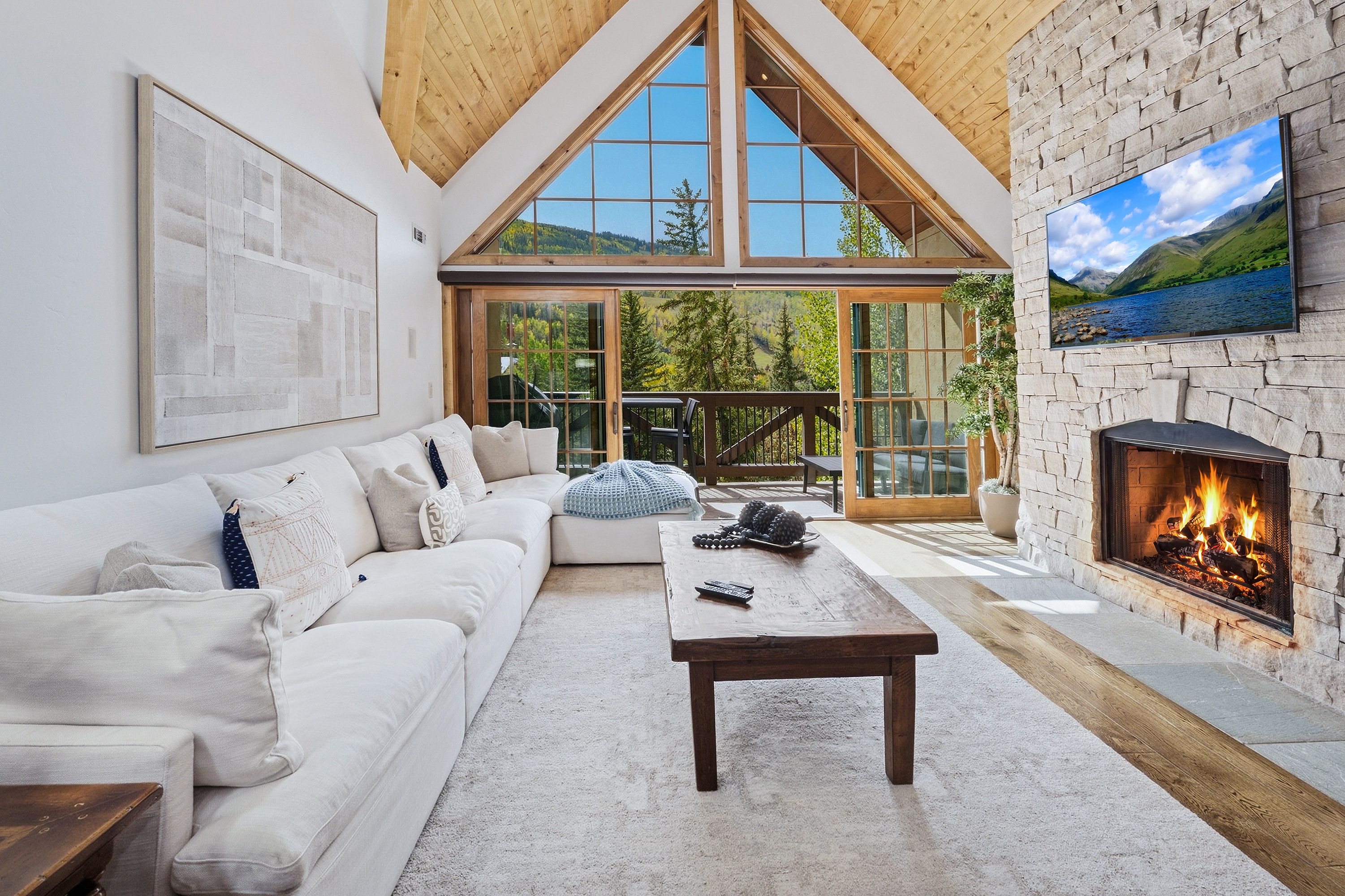 A cozy living room with a white sofa, a wooden coffee table, a fireplace, and a large window overlooking a mountainous landscape.