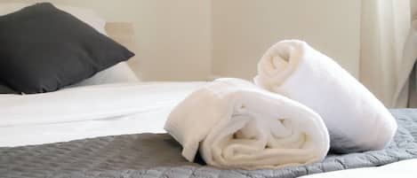 Una cama con colcha gris acolchada, una almohada blanca y dos toallas enrolladas en blanco.