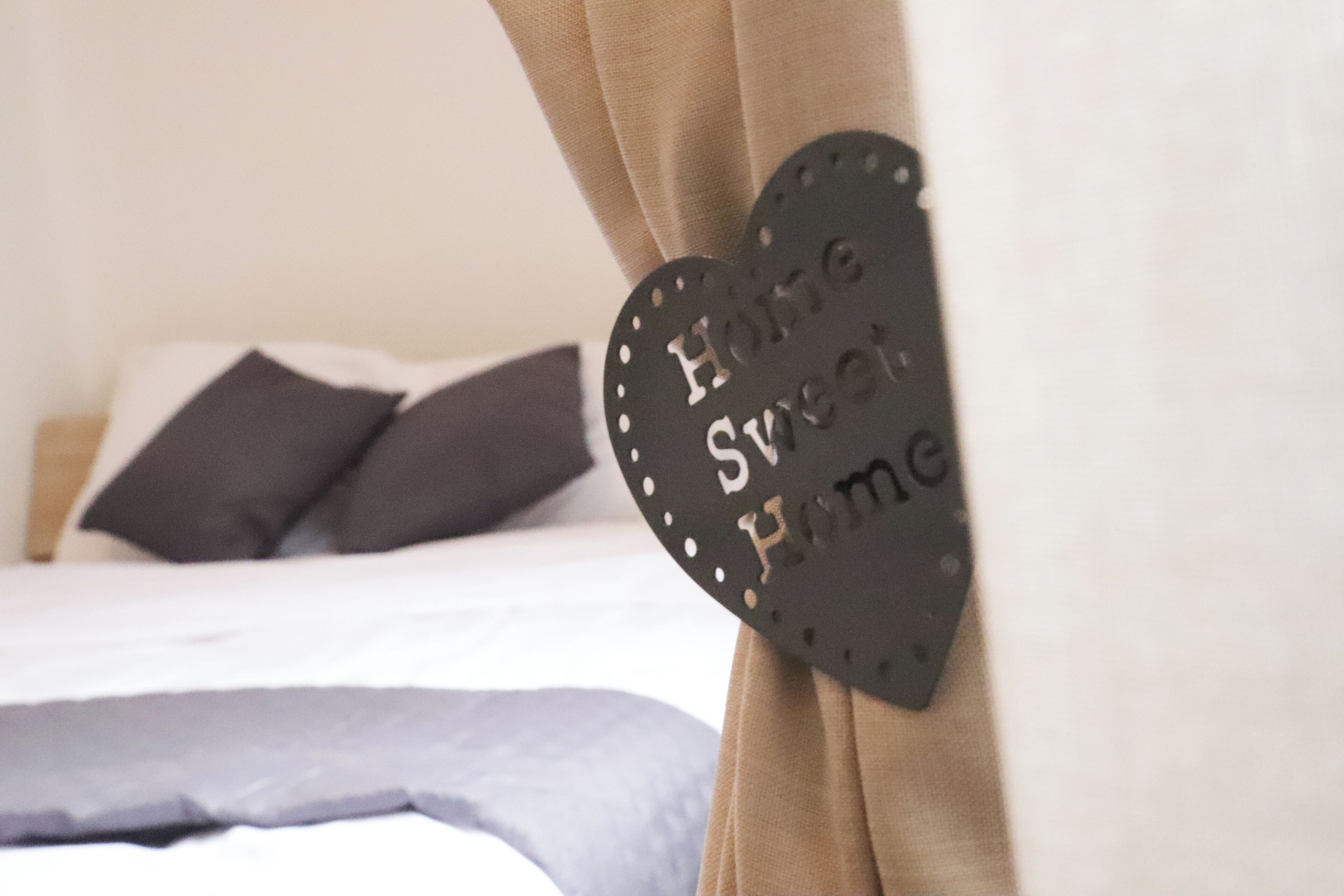 Una cama con una paleta de colores blanco y gris, una decoración con forma de corazón y el texto 'Home Sweet Home', y una cortina al fondo.