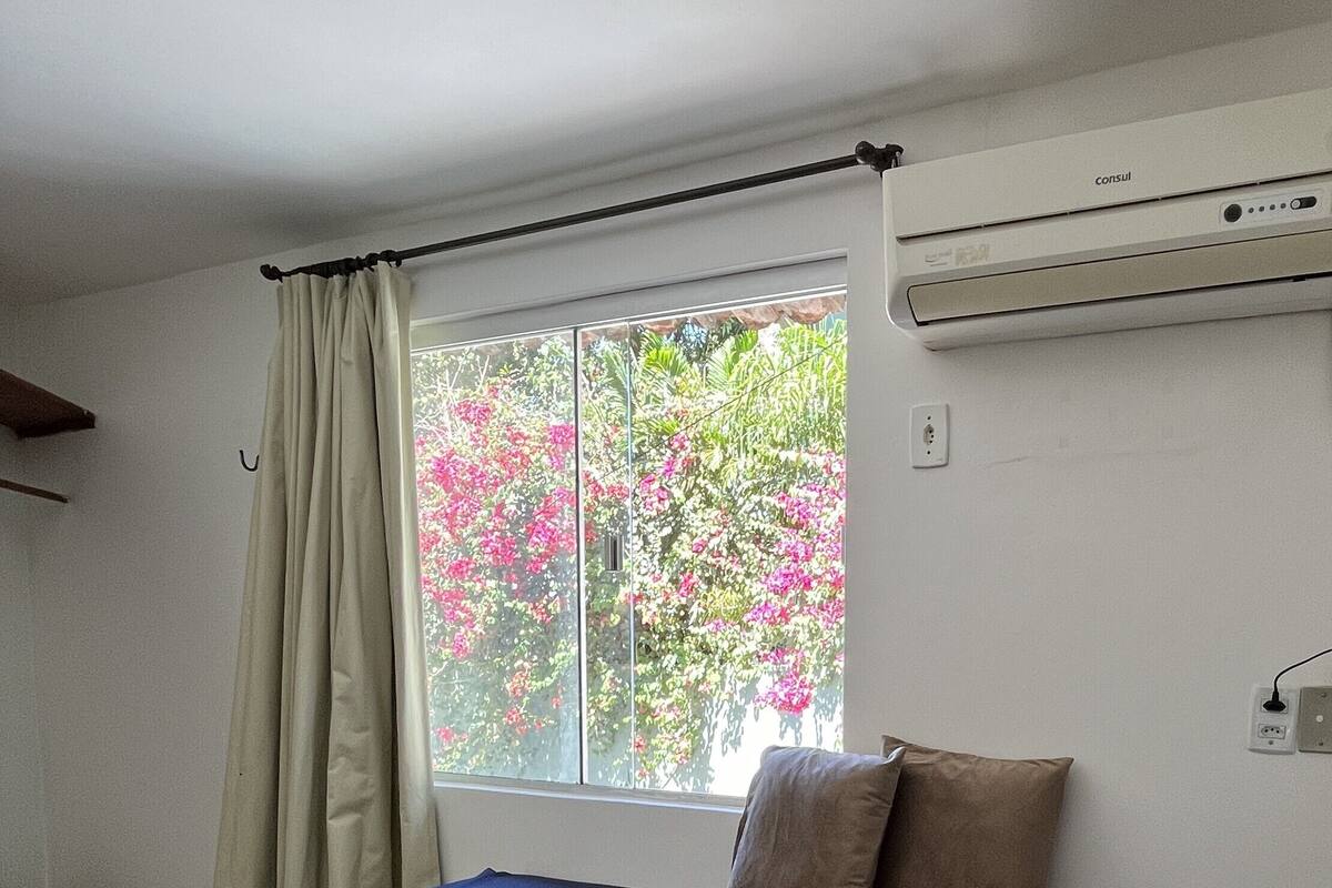A bedroom with a single bed, a window with curtains, an air conditioning unit, and a view of pink flowers outside.