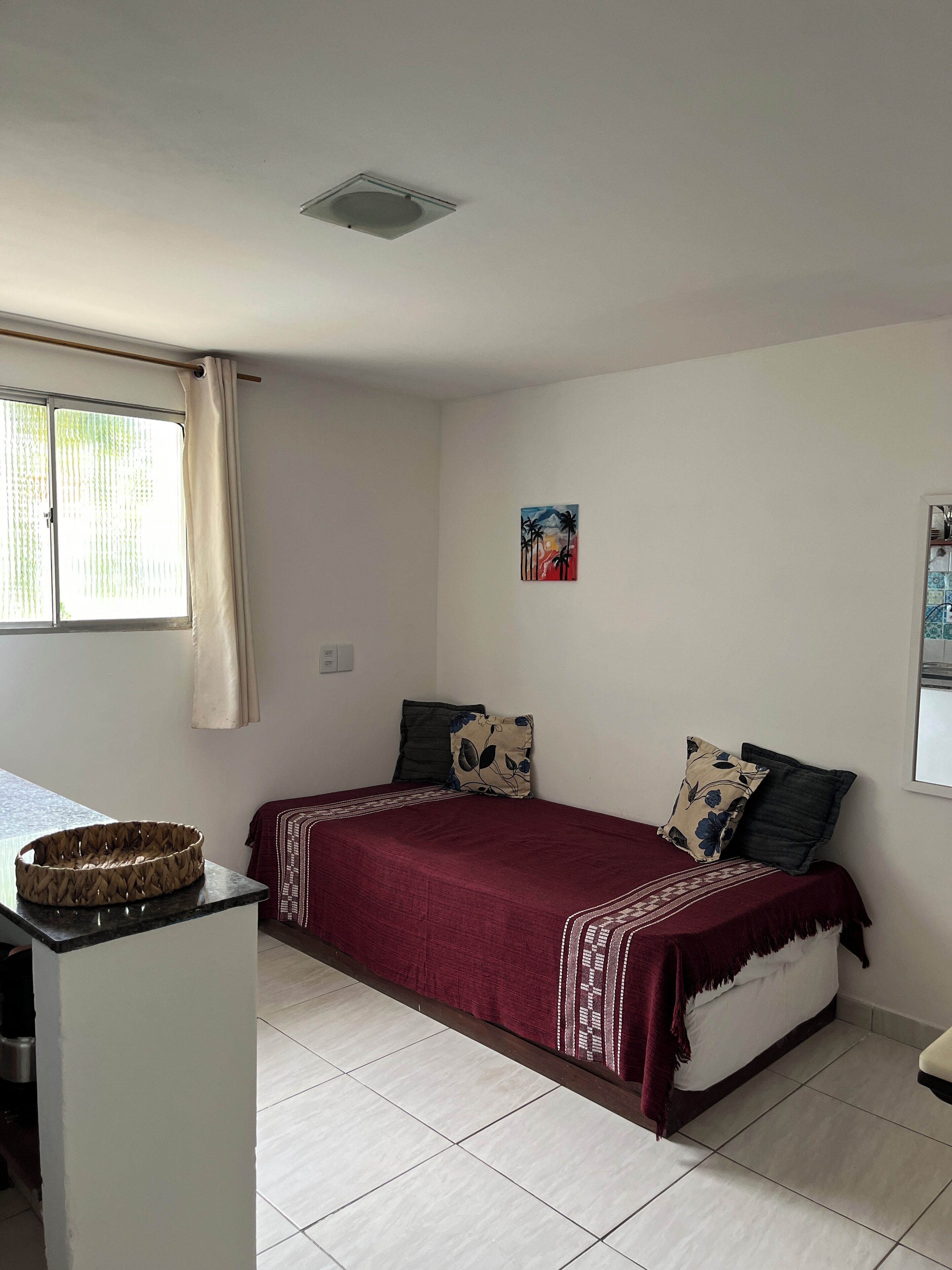 A single-bed room with a window, a ceiling light, a wall-mounted artwork, and a small table with a basket.