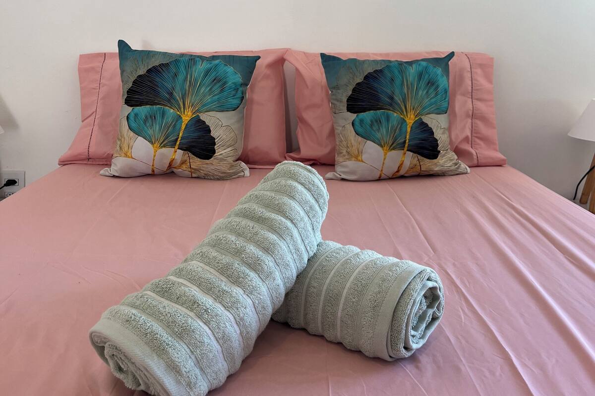 A neatly made bed with pink bedding, two decorative pillows with a leaf pattern, and a framed artwork on the wall.
