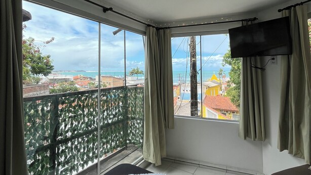 A room with a bed, a TV, and a balcony with a view of buildings and the sea. - Cores da Pipa flats (Tibau do Sul)