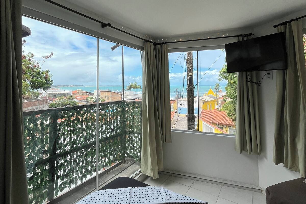 A room with a bed, a TV, and a balcony with a view of buildings and the sea.