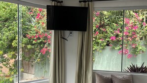 A bedroom with a large window, a bed, a flat-screen TV, and a view of a garden with pink flowers. - Cores da Pipa flats (Tibau do Sul)