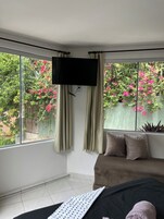 A bedroom with a large window, a bed, a flat-screen TV, and a view of a garden with pink flowers.