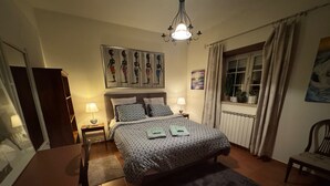A bedroom with a bed, two bedside tables, a chair, a desk, a window with curtains, a radiator, and a painting on the wall. - Casa Vimeiro Sleeps 2 With Pets (Alcobaça)
