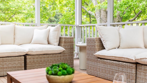 A wicker sofa with white cushions, a wooden table with a bowl of green plants, and a glass of water.