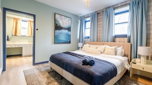A bedroom with a large bed, a bedside table, a nightstand, a window with curtains, a painting on the wall, and a bathroom with a mirror and towel rack.