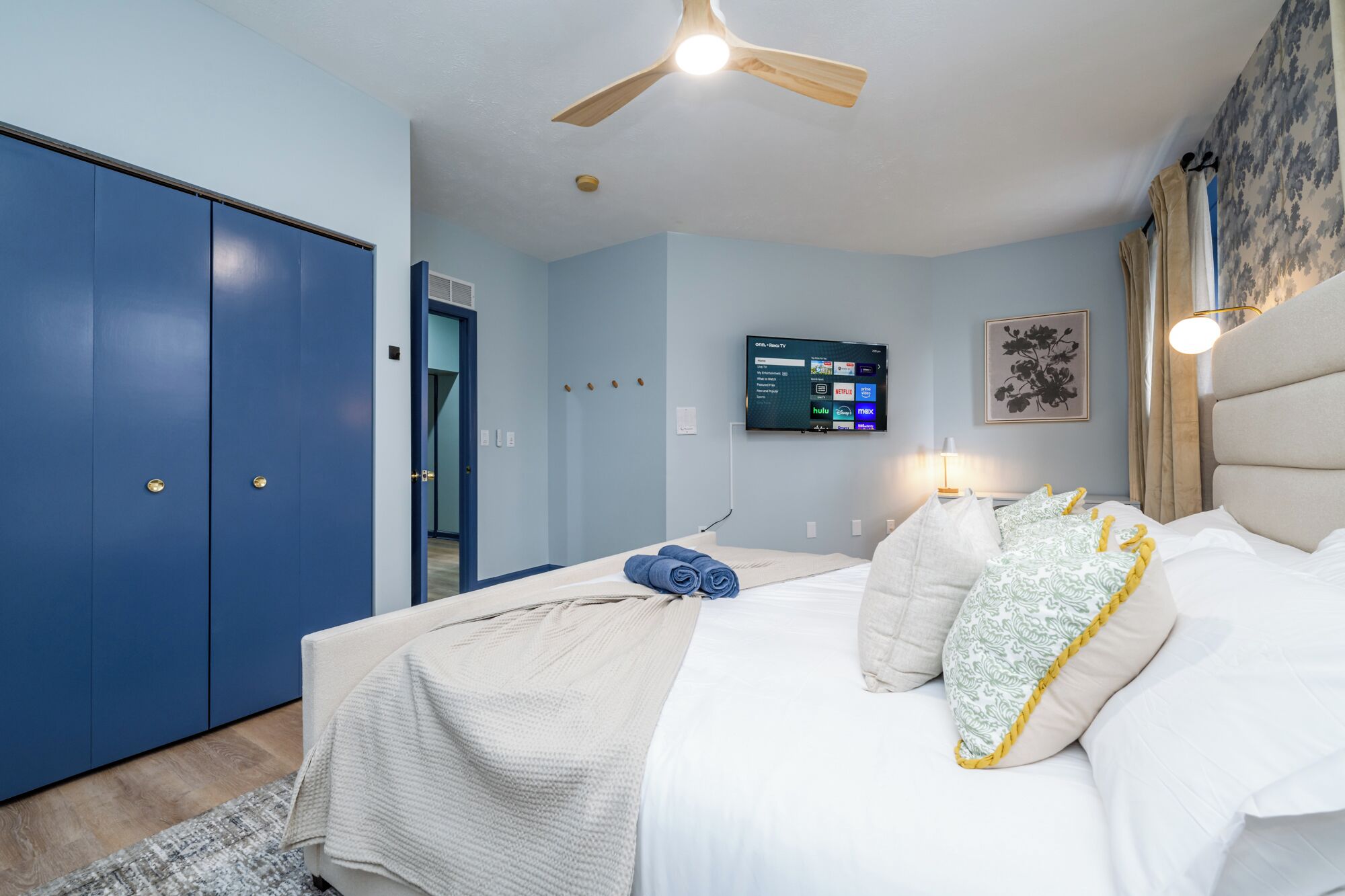 A bedroom with a bed, a ceiling fan, a TV on the wall, and a blue wardrobe.