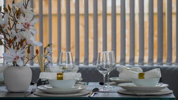 Mesa de comedor dispuesta con platos, tazones y vasos blancos, junto con un jarrón de flores blancas.