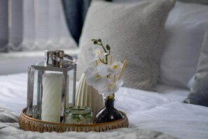 Una mesita de noche con una vela, un tarro de cristal y flores blancas sobre la cama.