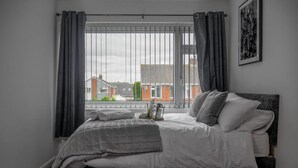 Un dormitorio con cama, cortinas grises, vistas a la ciudad y un cuadro en la pared.