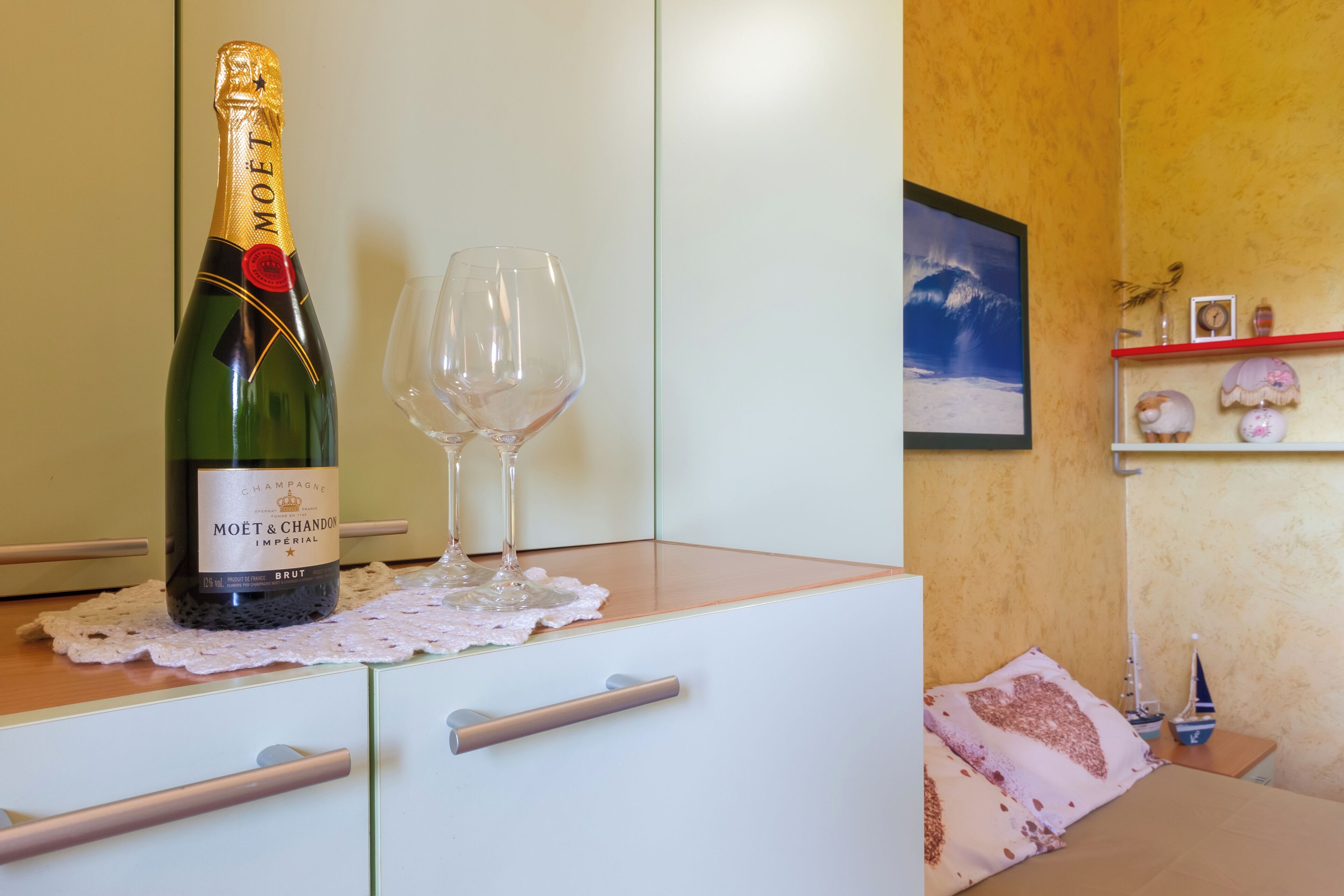 A bottle of Moët & Chandon champagne and two glasses on a table in a room with a bed, a shelf with decorative items, and a framed picture on the wall.