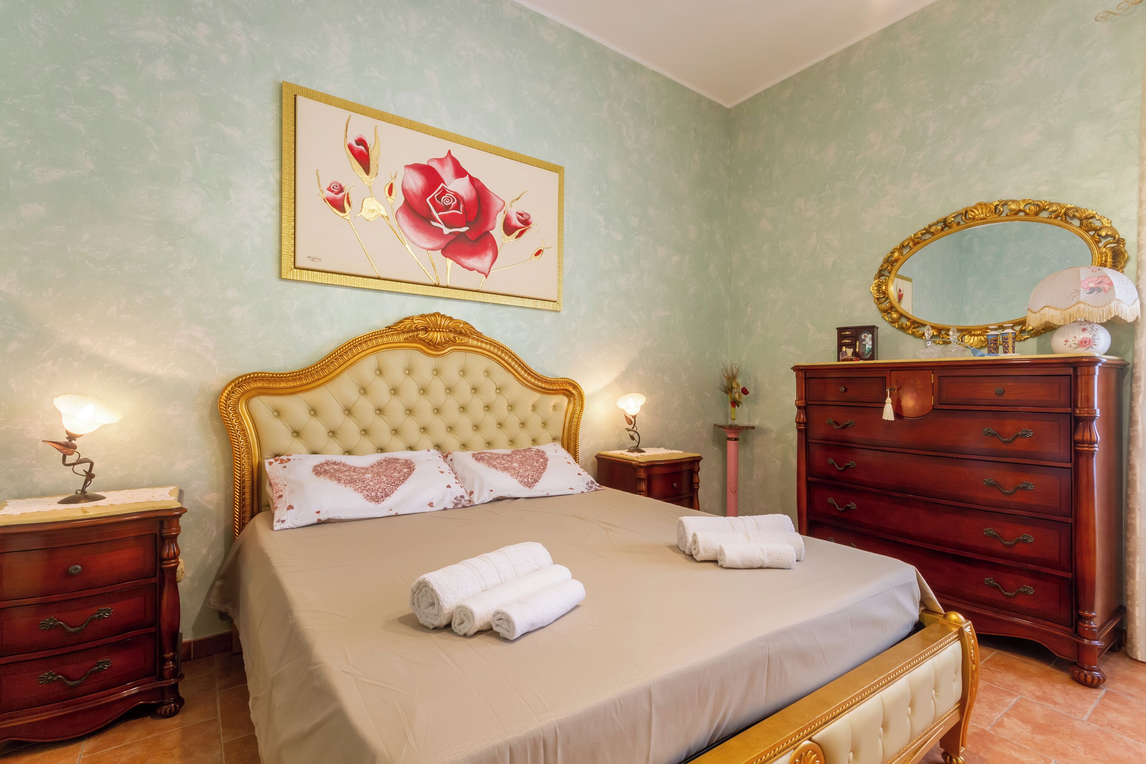 Una cama bien hecha con cabecera, una mesita de noche con lámpara, un aparador con espejo y una obra de arte floral enmarcada en la pared.