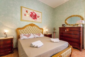 A neatly made bed with a headboard, a bedside table with a lamp, a dresser with a mirror, and a framed floral artwork on the wall.