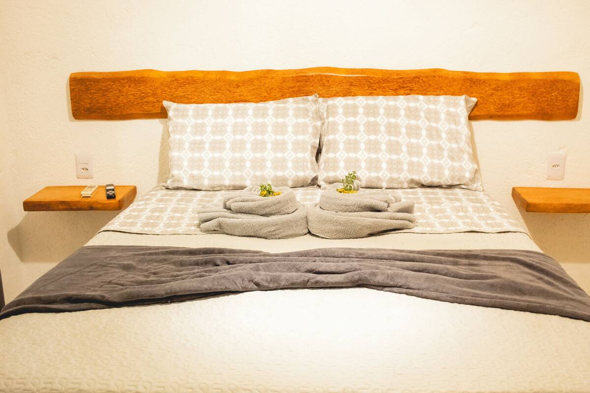 Una cama bien hecha con un cabecero de madera, dos almohadas con un estampado geométrico y dos mesitas de noche con controles remotos.