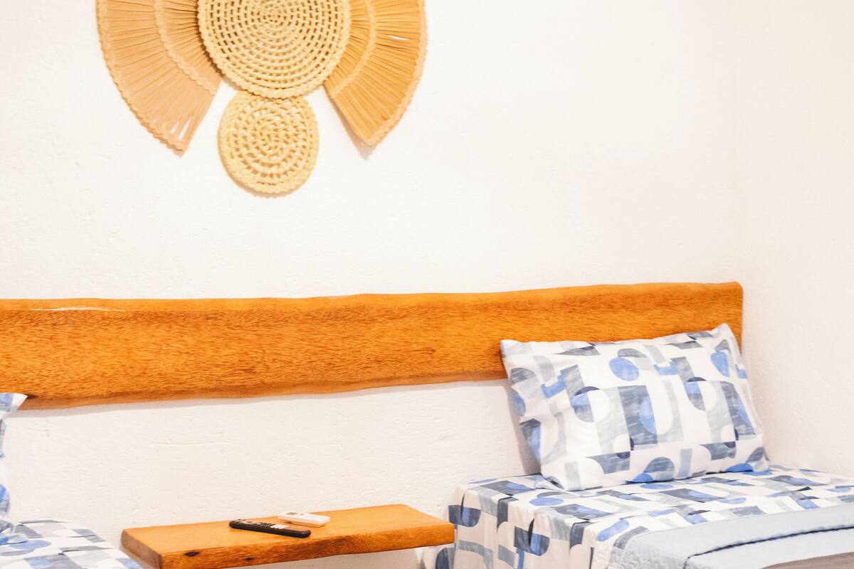 Un dormitorio con dos camas, cabecero de madera, ropa de cama estampada, un adorno mural tejido y un pequeño estante de madera.