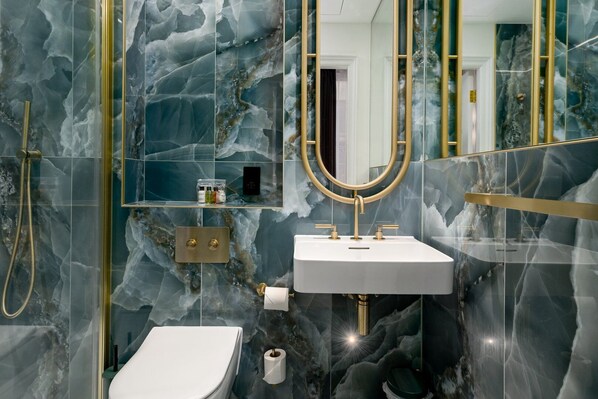 A modern bathroom with marble walls, a gold-framed mirror, a white sink, and a shower.