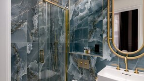 Een badkamer met een douche met glazen wand, marmeren wanden, een spiegel met gouden lijst en een wit wastafelmeubel met gouden details.