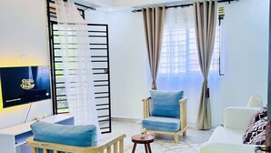 A modern living room with a flat-screen TV, two blue cushioned chairs, a white sofa, a wooden coffee table, and a ceiling light fixture. - Apartment unit-Pearl Residence (Kampala)