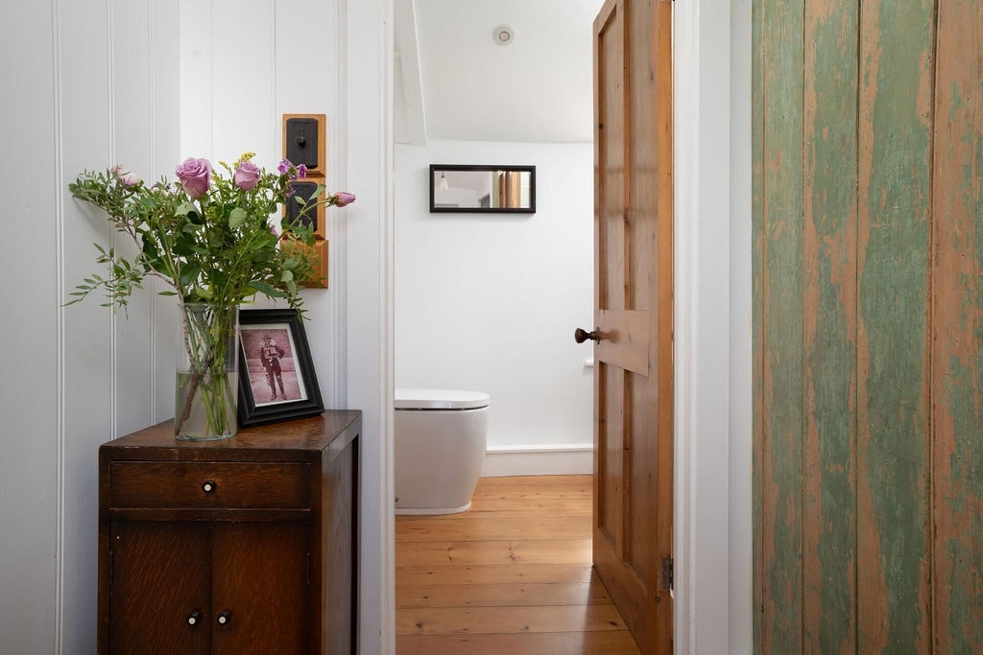 A hallway with a wooden door, a white toilet, a framed photo on a small wooden cabinet, and a vase with flowers.