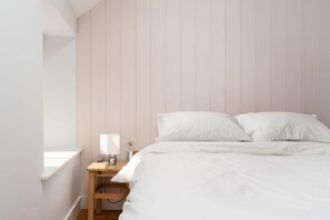 Et soveværelse med seng, hvide sengetøj, et natbord med lampe og et vindue.