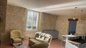 A room with a sofa, armchair, and a small table with a remote control. There is a window with blinds, a stone wall, and a kitchenette area with a sink and countertop. - House Air conditioning in the countryside Vouvray (Vouvray)