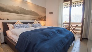 A bedroom with a large bed, a nightstand, a wall-mounted lamp, a painting of a mountain landscape, and a balcony with a table and chairs.