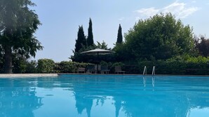 A clear blue swimming pool with lounge chairs and umbrellas, surrounded by trees and a clear sky.