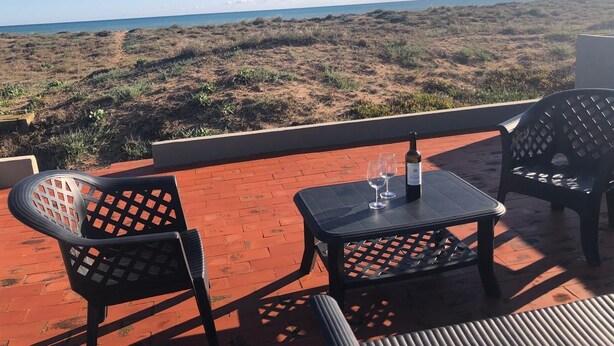 Una terraza con dos sillas y una mesa, con vista a una playa con dunas y el océano.