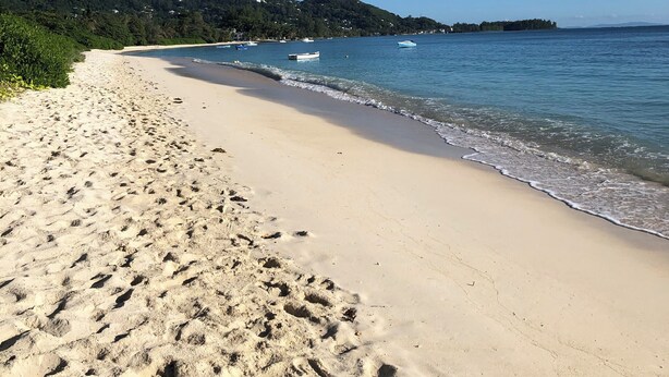 Platja sorrenca amb empremtes, cel blau clar i mar tranquil·la amb vaixells.