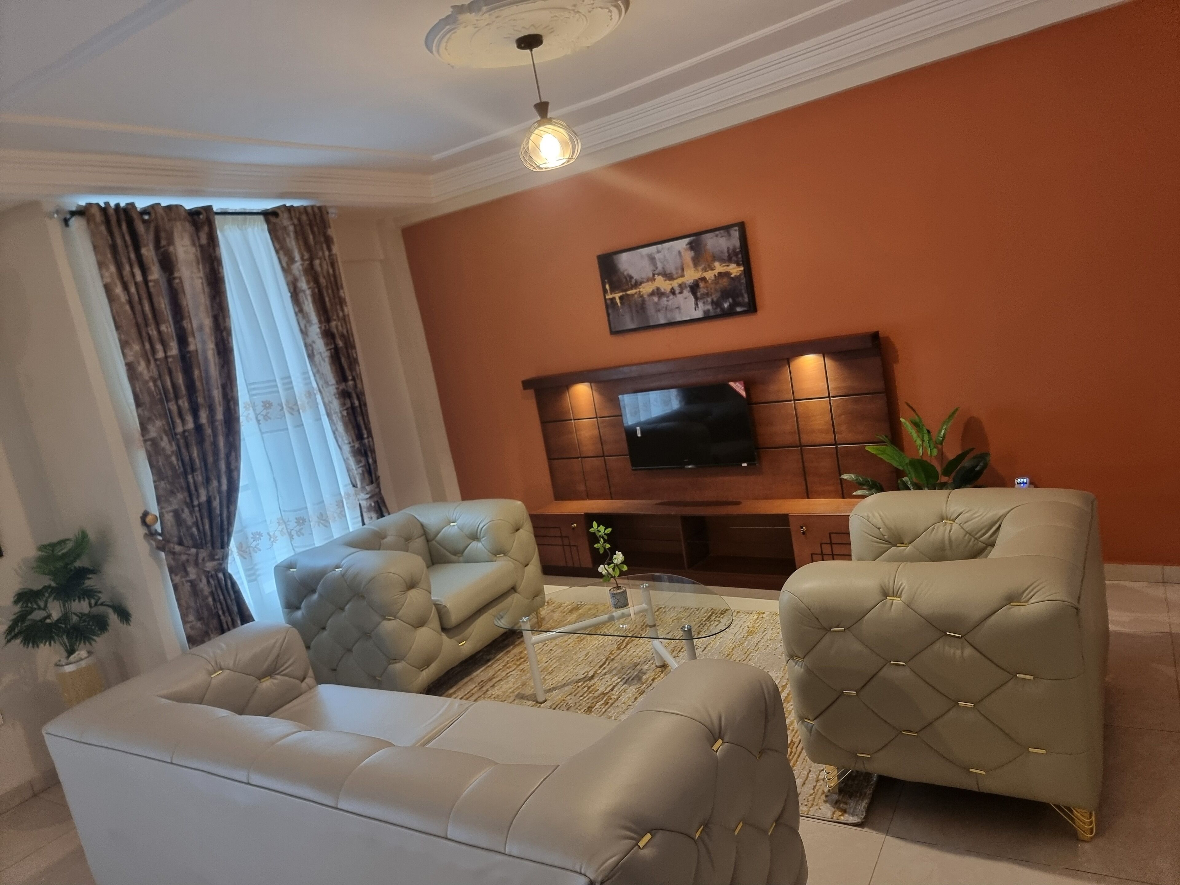 A living room with a sofa, two armchairs, a coffee table, a TV mounted on a wooden cabinet, and a ceiling light.