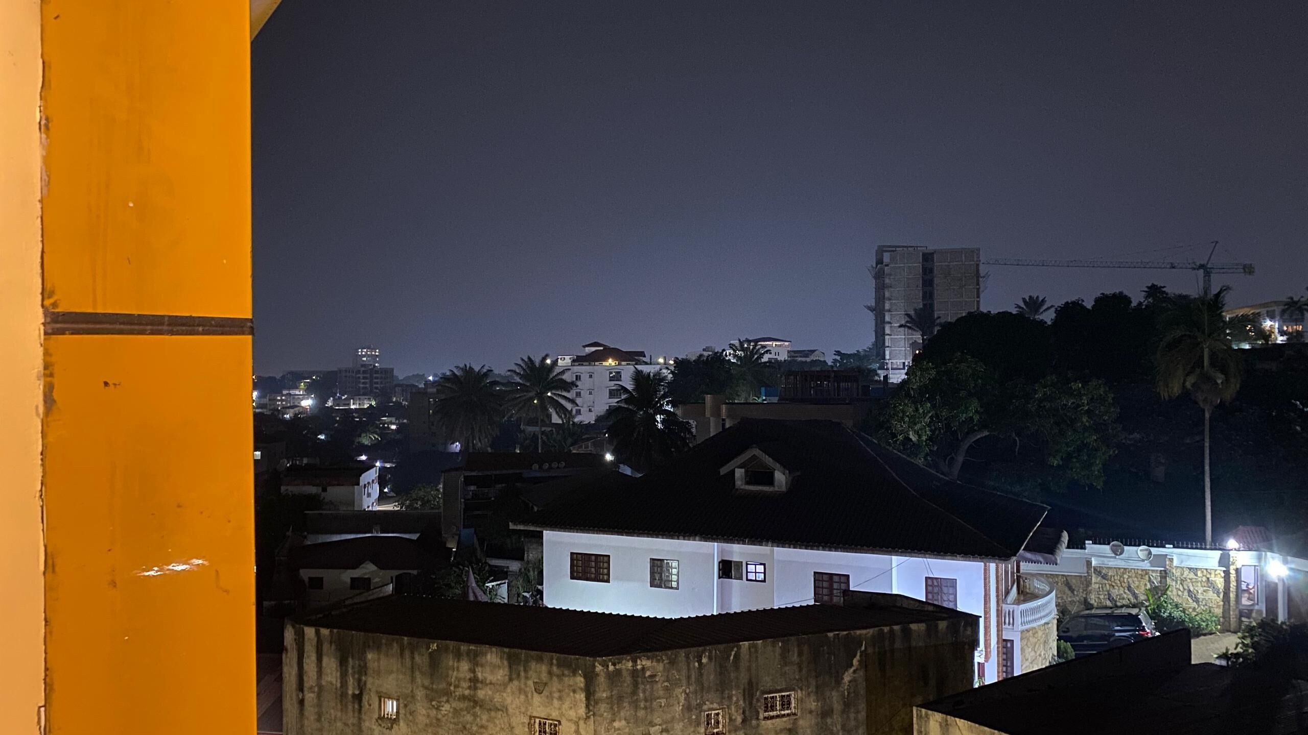 A nighttime view of a cityscape with buildings, including a white house with a dark roof, under construction, and a crane in the background.
