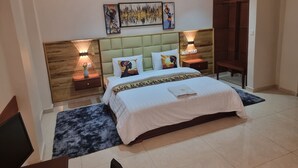 A modern hotel room with a large bed, two bedside tables with lamps, a headboard with artwork, and a wooden wardrobe. - Résidence hôtelière MEDOM Palace, Bastos (Yaoundé)