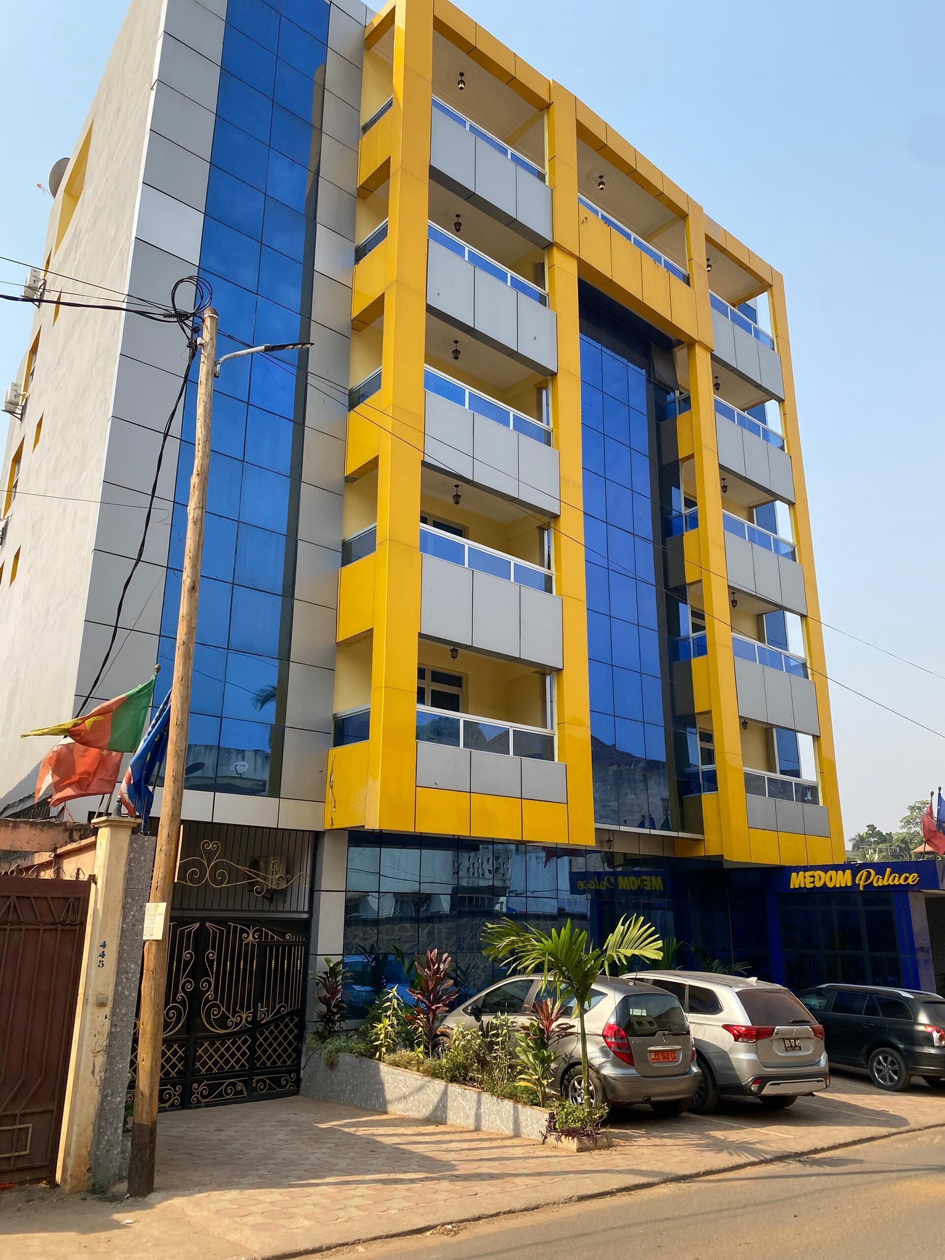 Un immeuble à plusieurs étages, doté d’une façade jaune et bleue, d’une entrée fermée par un portail et d’une enseigne indiquant « Palais MEDOM ».