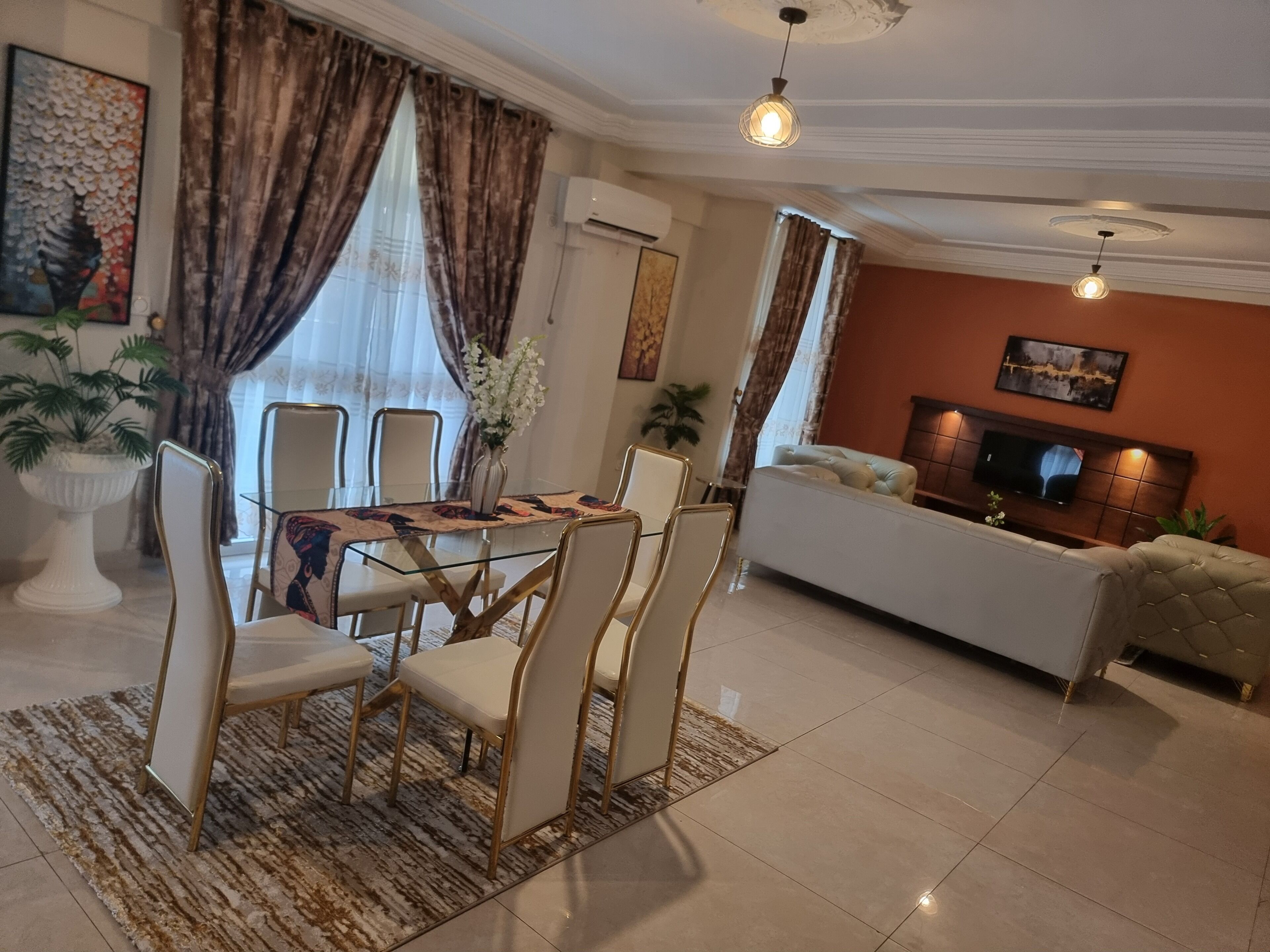 A modern living room with a dining area, featuring a glass-top table, white chairs, a sofa, and a TV.